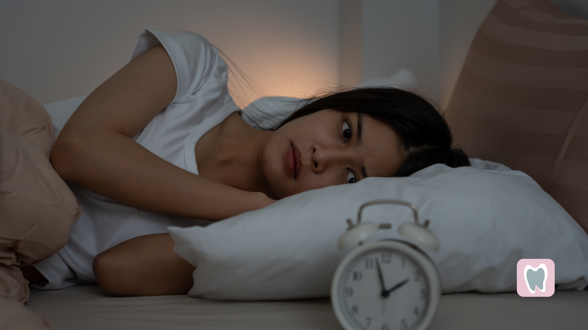 Woman lying in bed on her side beside a white alarm clock in a dimly lit bedroom