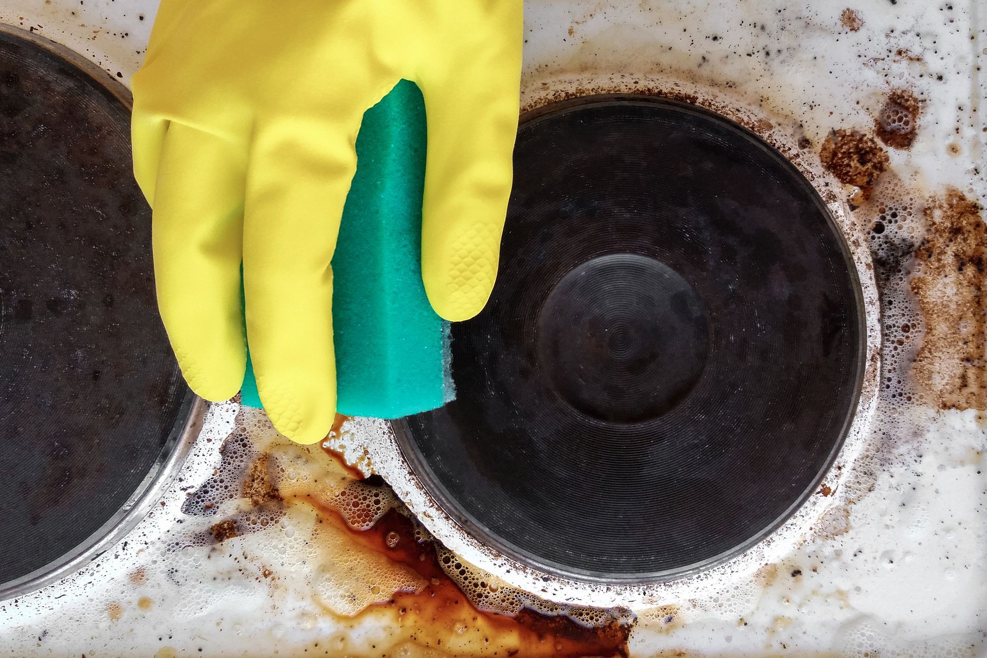 A person wearing yellow gloves is cleaning a stove top with a green sponge.