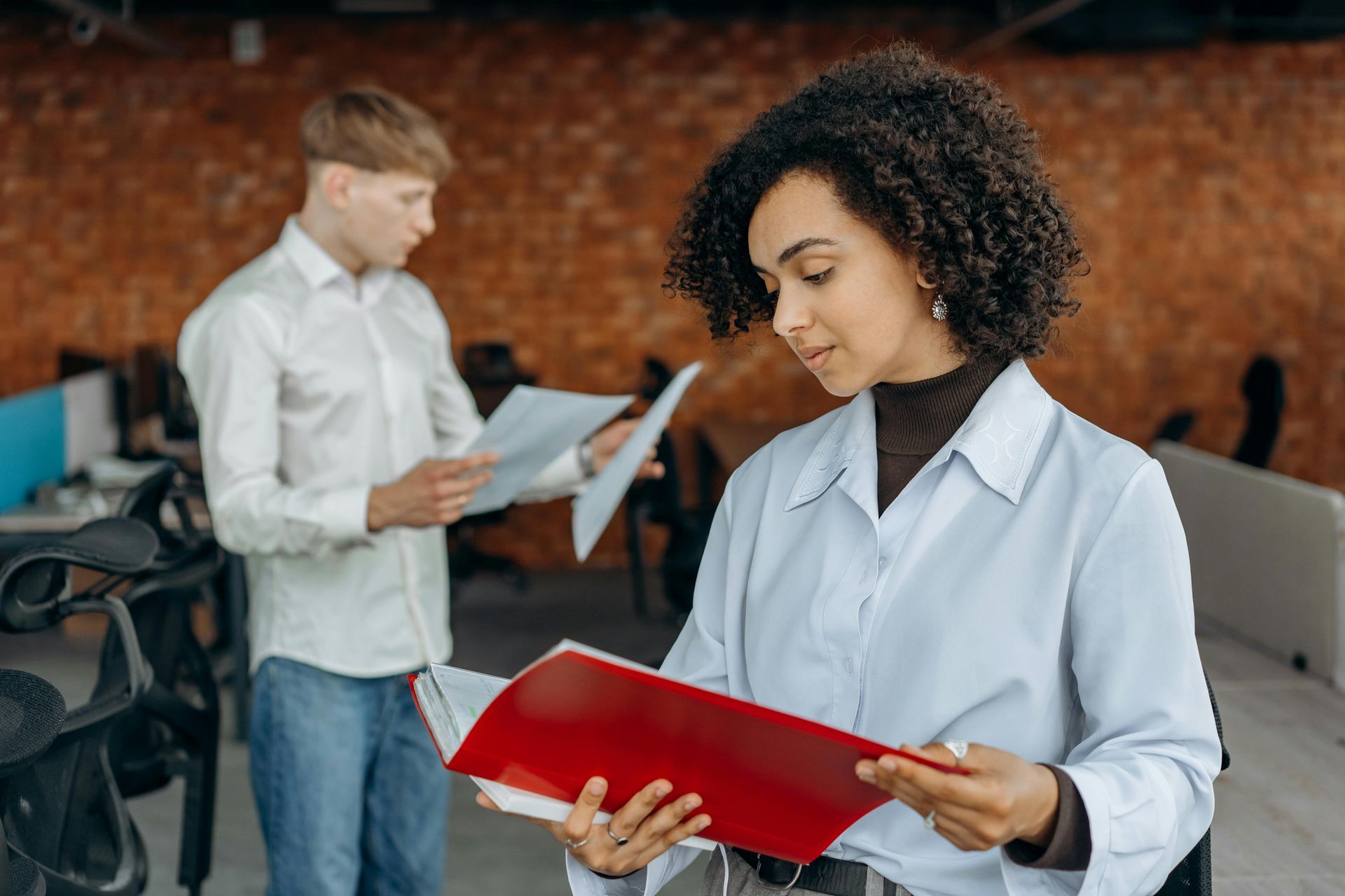 Woman with curly hair in a light blue shirt looks at a red binder. Man in white shirt and jeans looks at papers. Office setting.