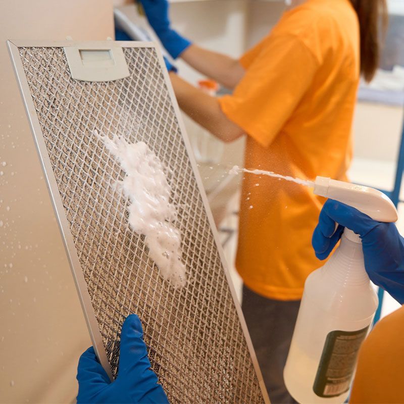 A woman is cleaning a range hood stove filter with a spray bottle.