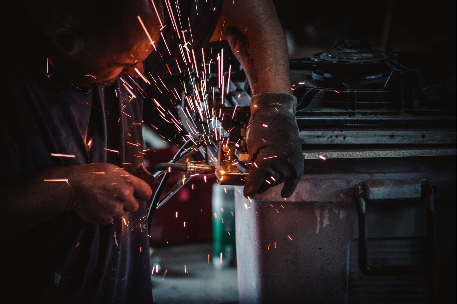 Worker welding metal, sparks flying in a dark workshop.