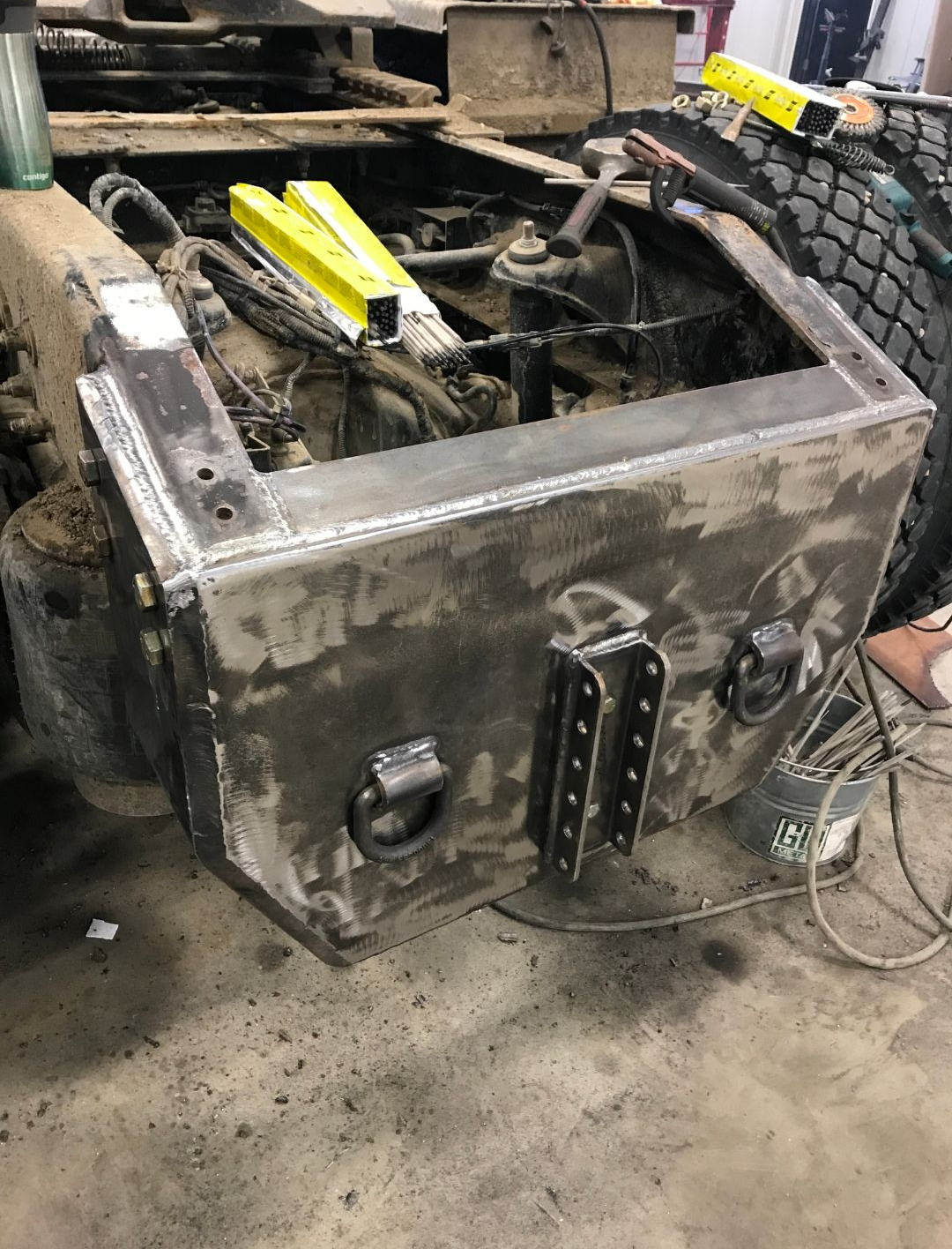 Large gray metal box or chassis in a workshop, with yellow parts and a tire visible inside.