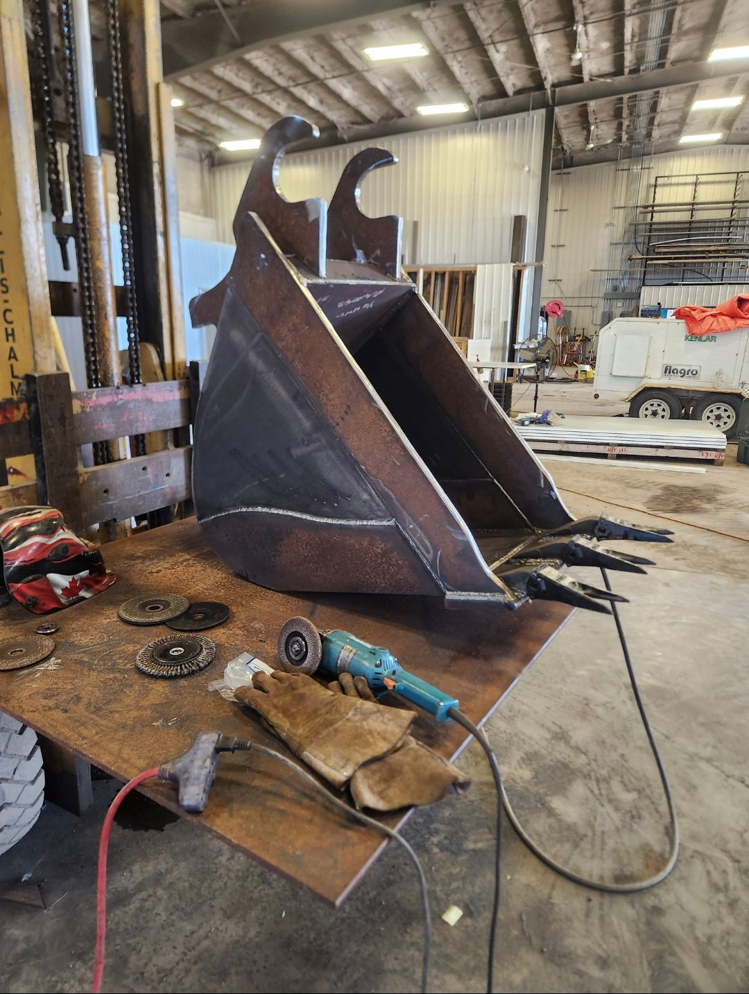 Large metal excavator bucket in a workshop, with tools and a welding cable on a workbench.