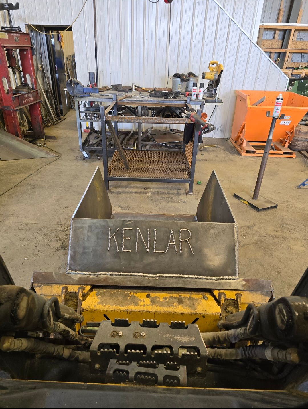 Factory floor with a yellow machine in front of a metal bin labeled “KENLAR” and workshop equipment behind.