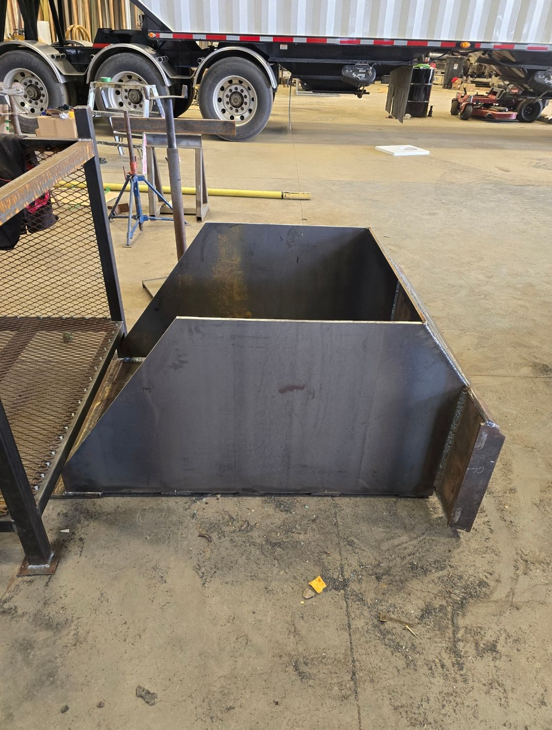Empty metal hopper on a concrete floor in an industrial yard, with a truck in the background