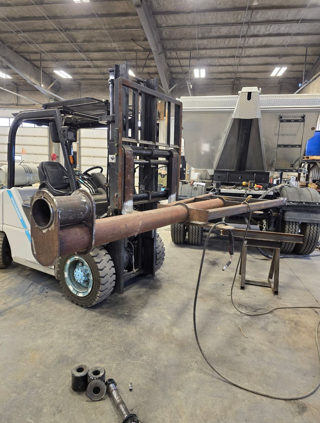 Forklift carrying a large rusted metal pipe in an industrial workshop, with tools and equipment nearby