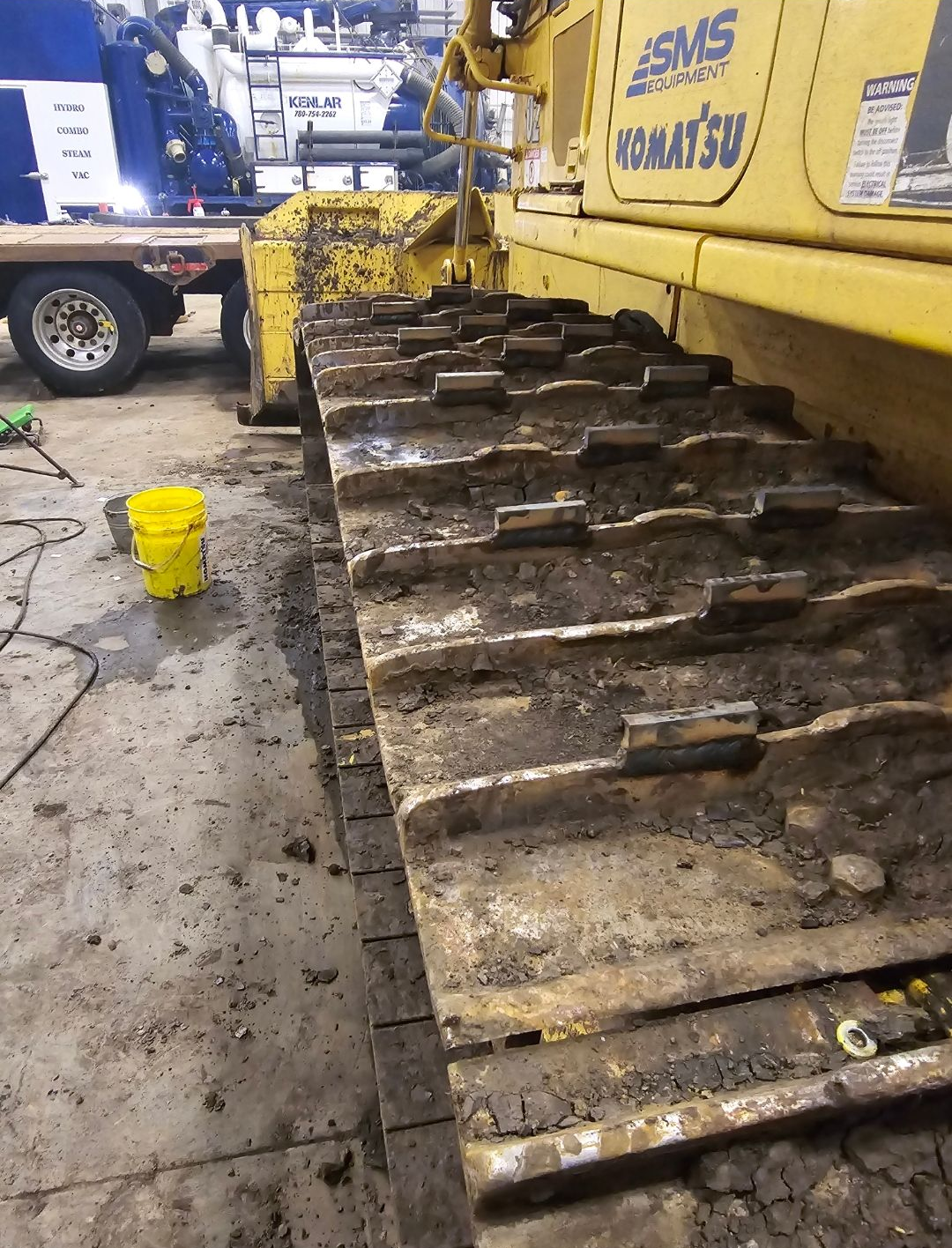Dirty yellow construction vehicle tracks on a worksite beside a white truck and yellow bucket