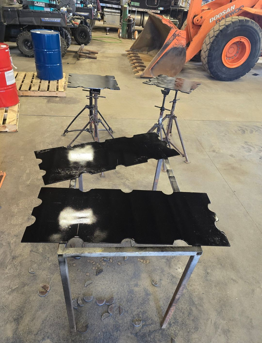 Black metal panels on a workbench in a workshop, with orange equipment and tripods in the background
