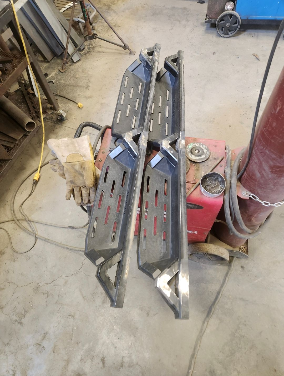 Three metal car side steps with red accents lying on a workshop floor