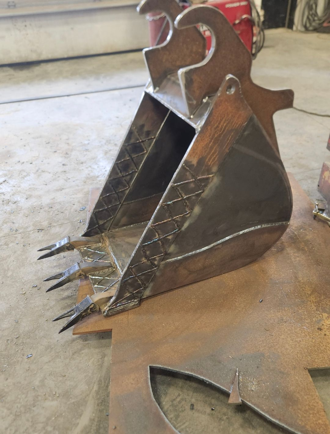 Steel excavator bucket with teeth resting on a concrete floor, close-up view