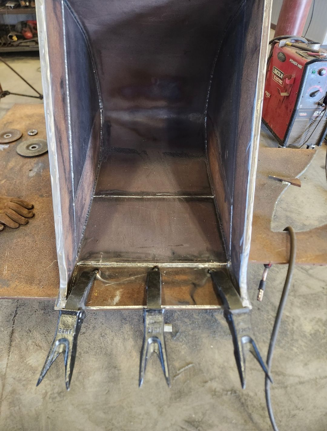 Metal chute with welded tines and brackets in a workshop setting, viewed from the open end.
