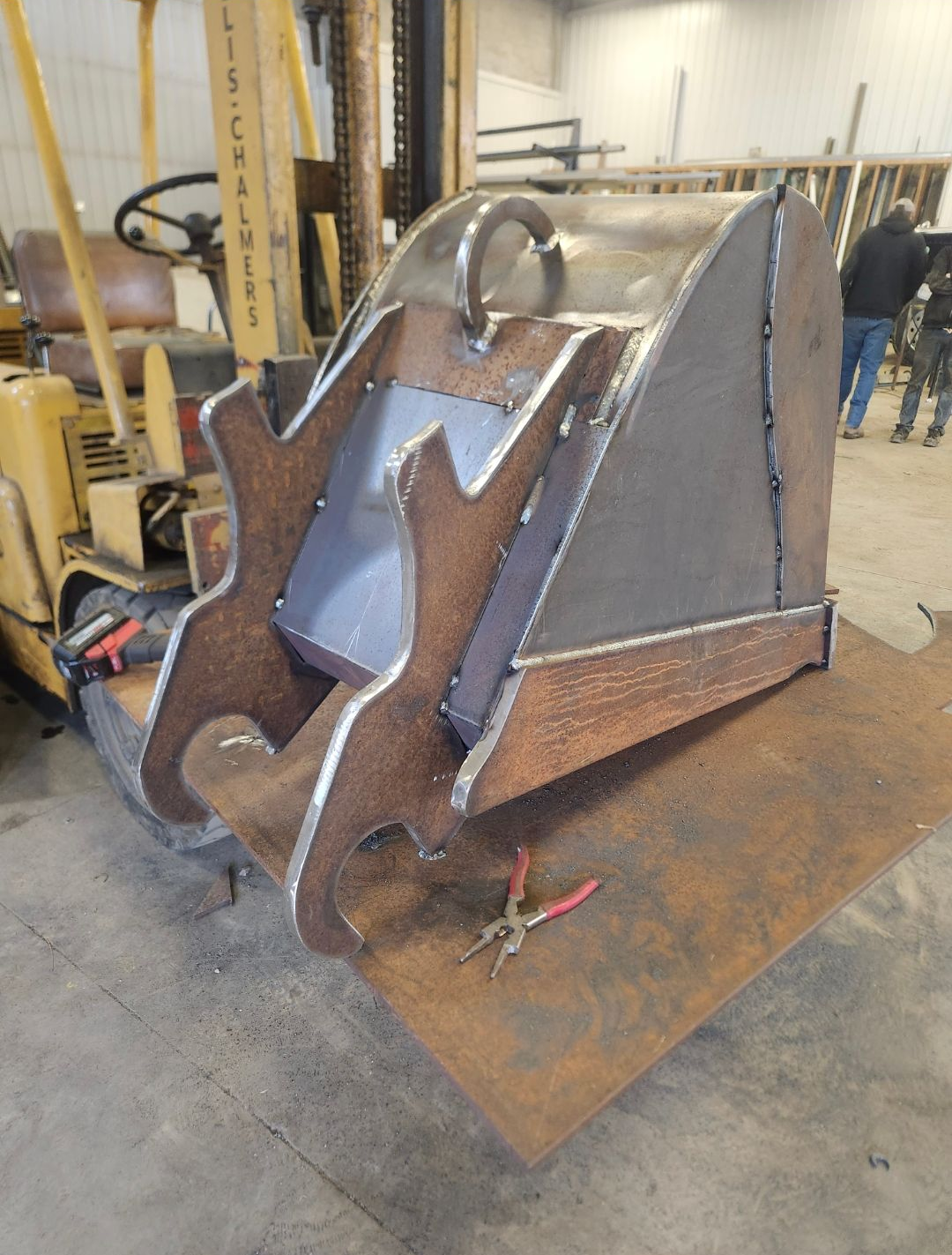 Steel bucket attachment in a warehouse, with welded edges and a rusted surface on a floor plate.