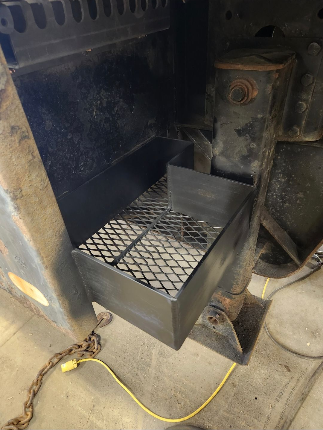 Black metal pull-out tray with wire mesh inside a large machine or industrial cabinet.