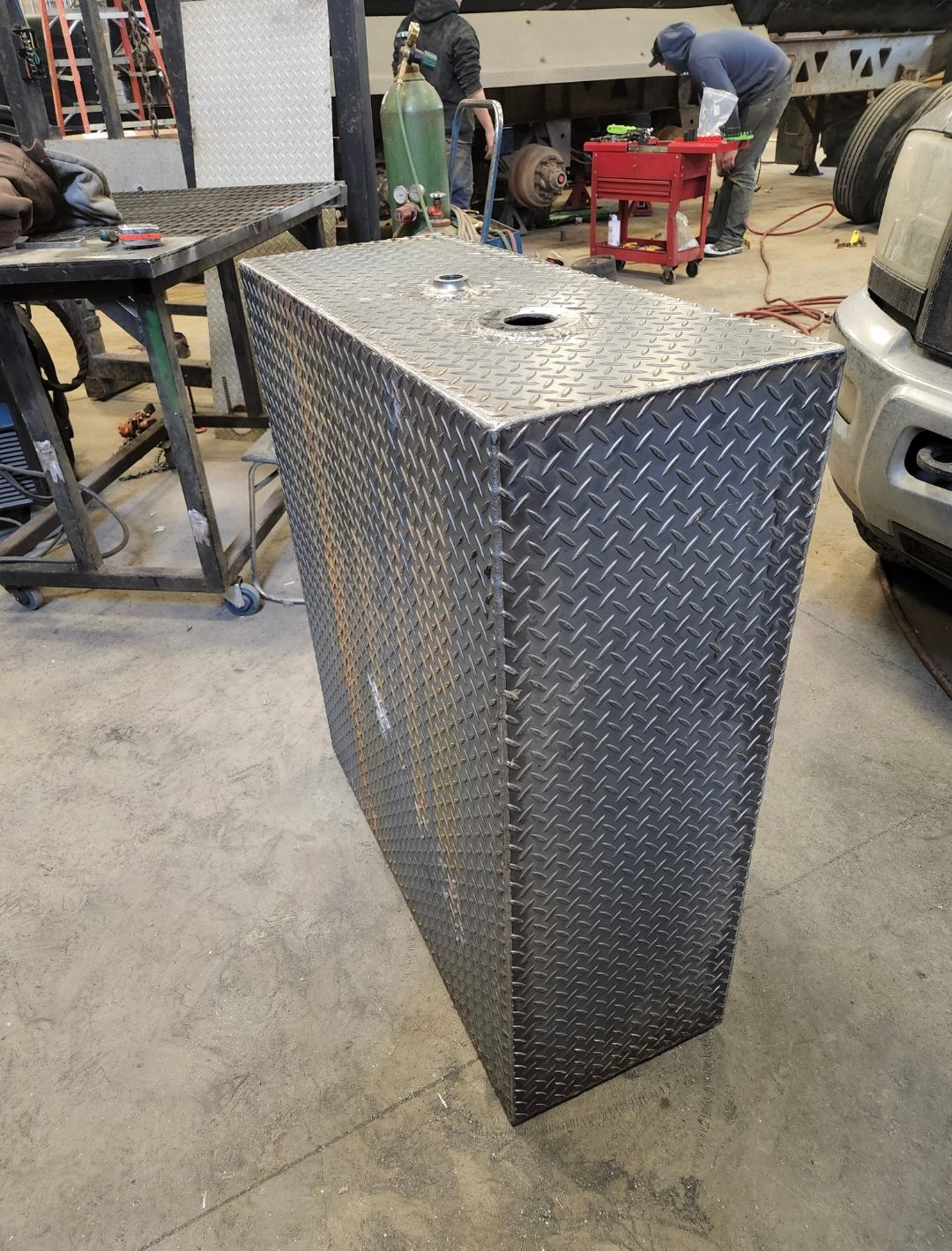 Tall diamond-plate metal storage box in a workshop garage, with tools and vehicles in the background