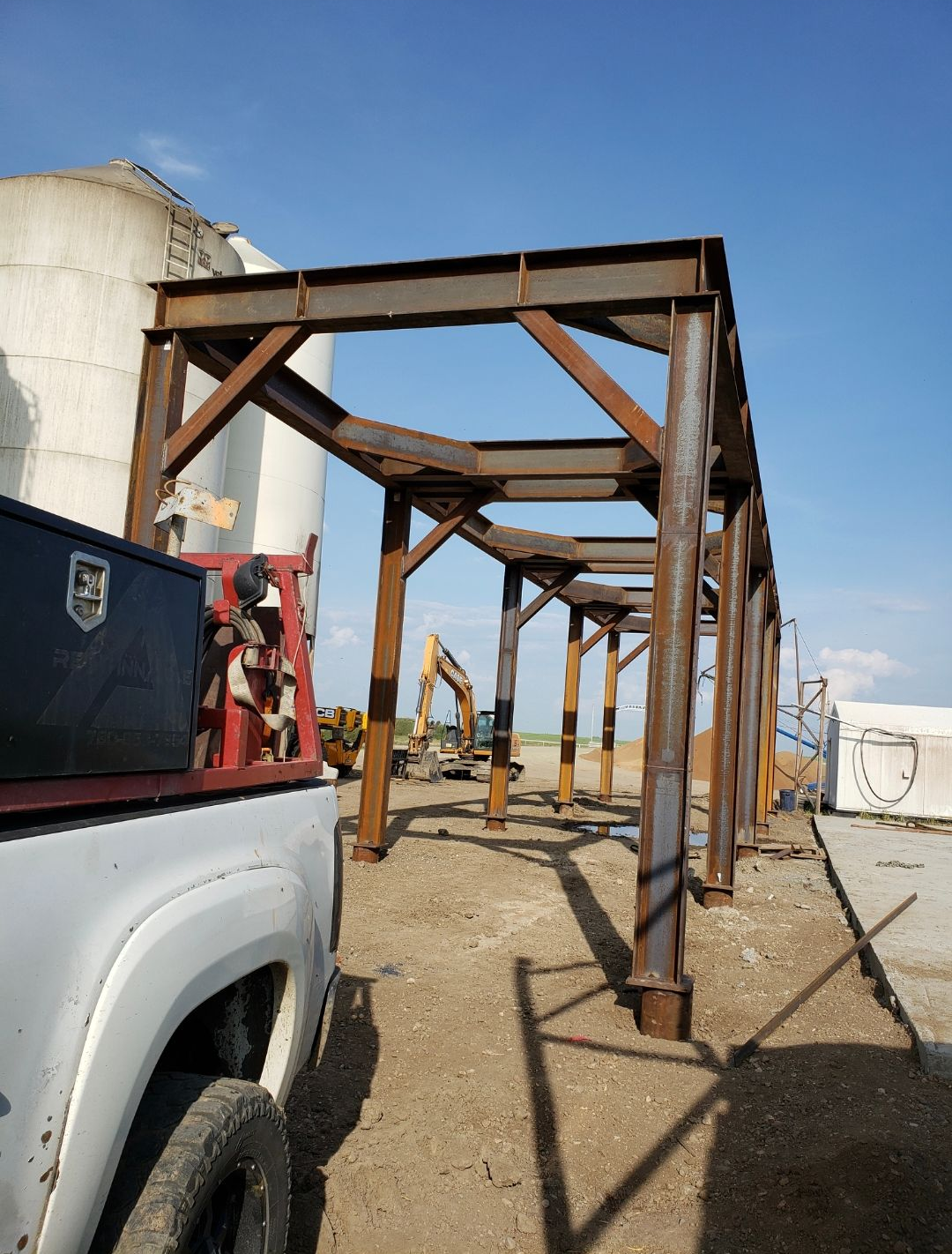 Rusty steel frame structure beside a white truck on a gravel lot under a blue sky.