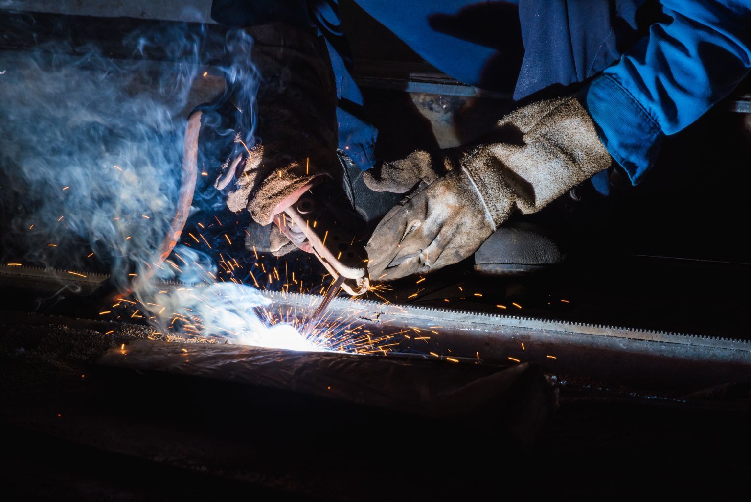 Gloved hands welding a metal pipe, with bright sparks and smoke in a dark setting