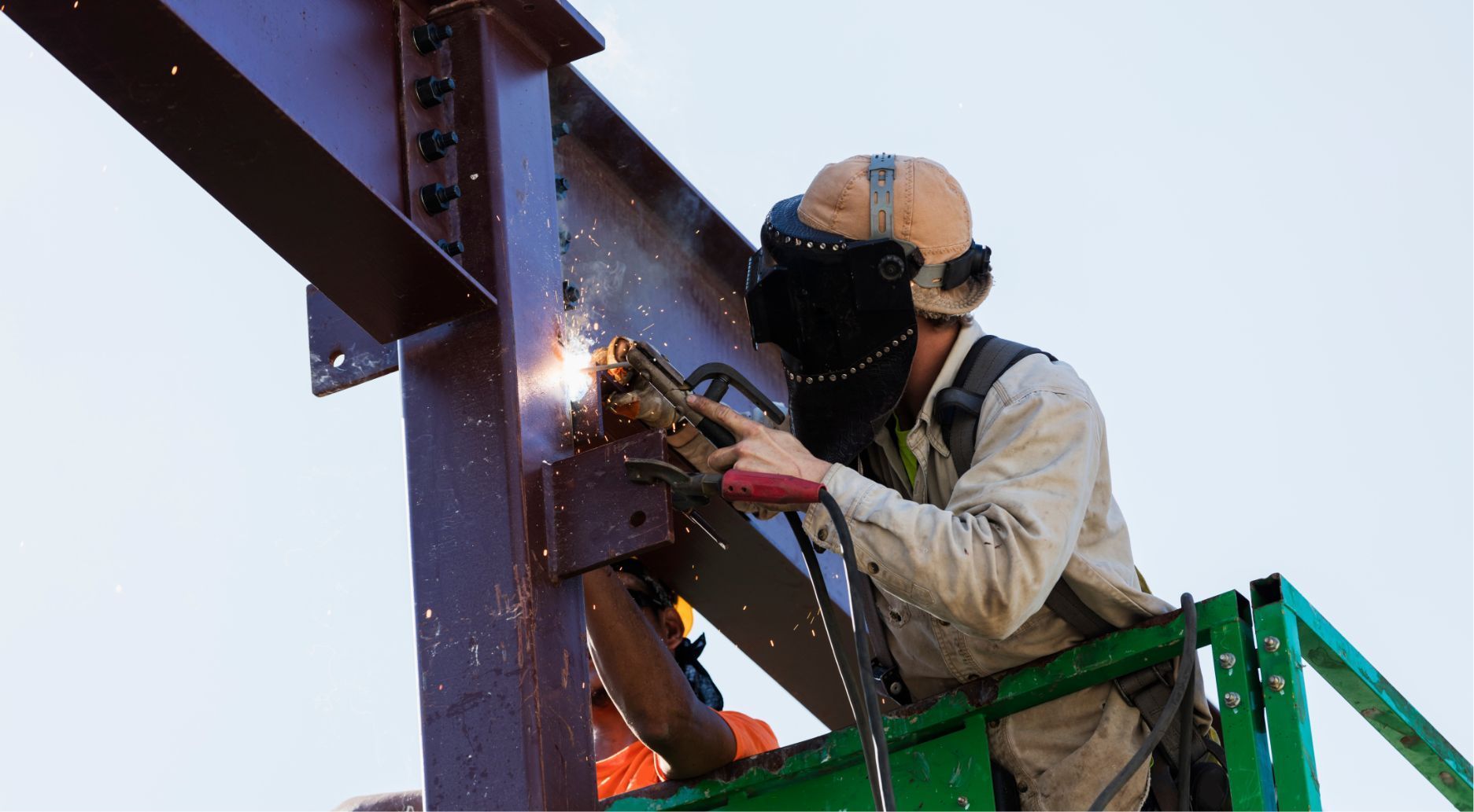 Worker welding a steel beam from a green lift, sparks flying, wearing protective gear