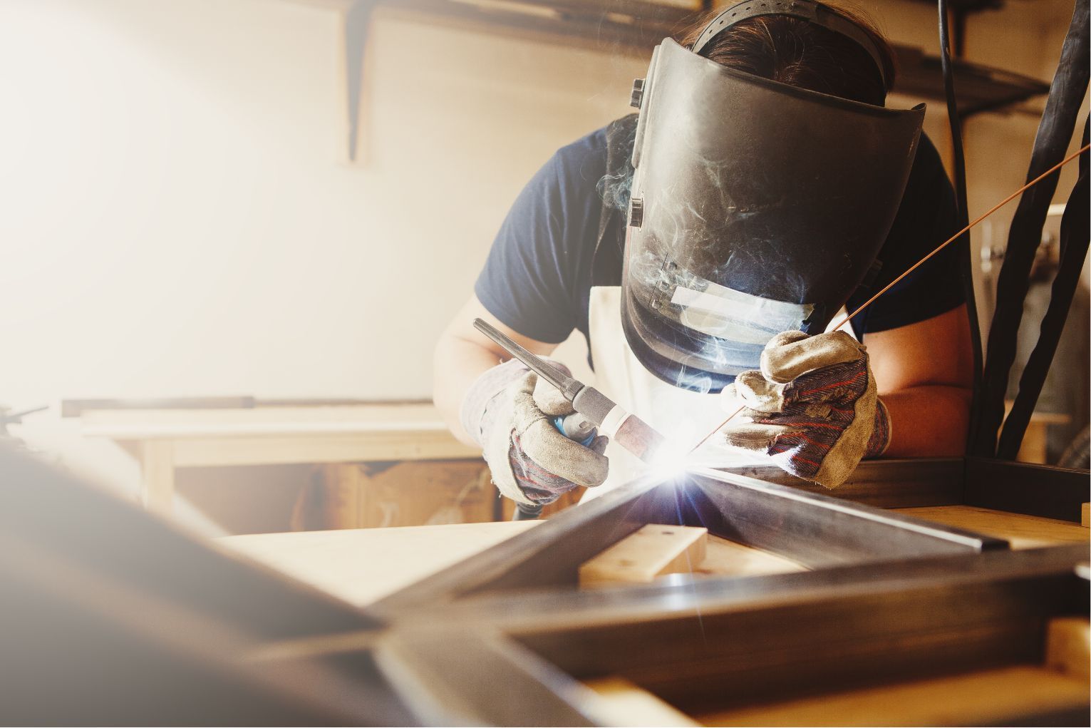 Person wearing a welding helmet works on a metal frame in a workshop.