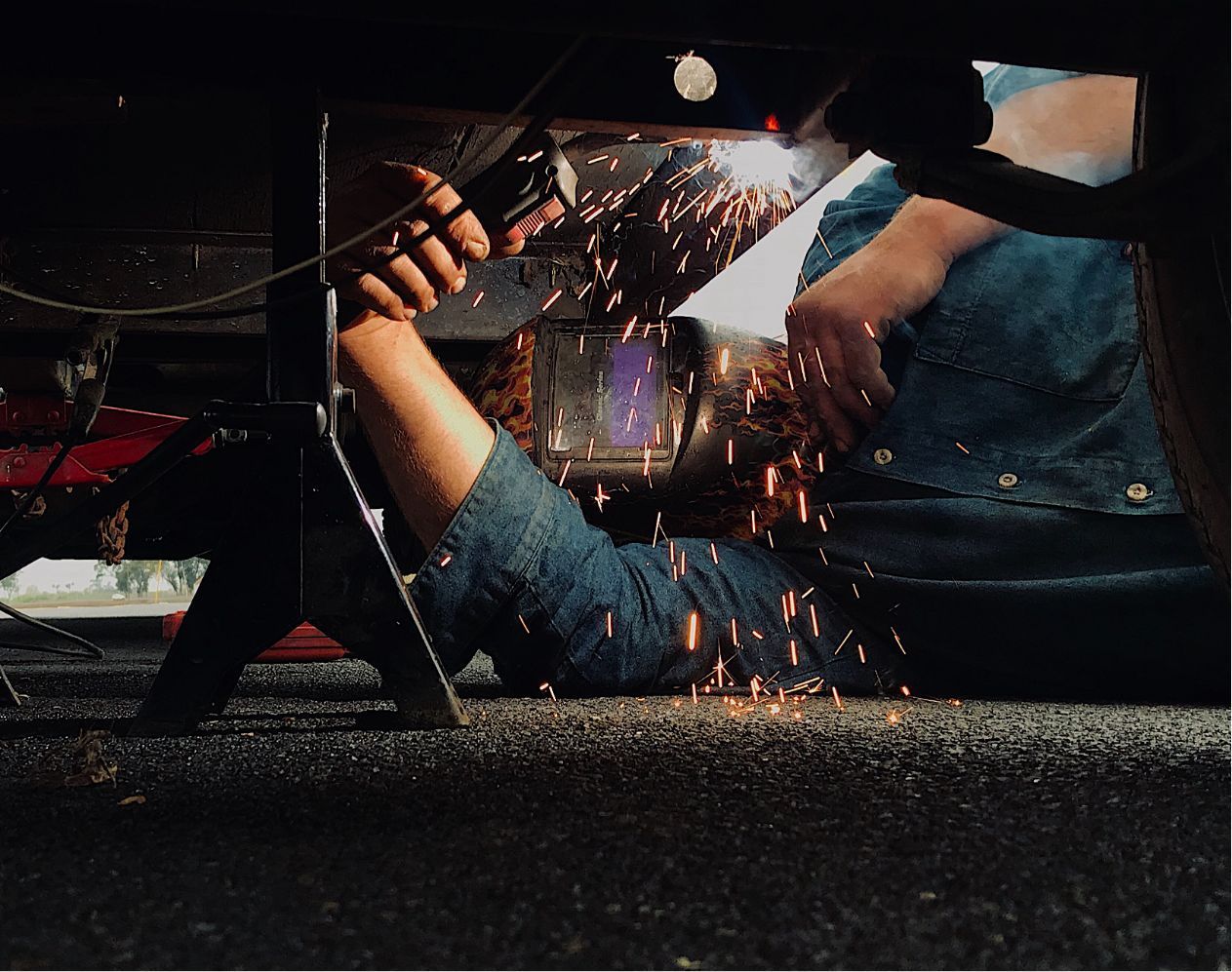 Welder in denim works under a vehicle, sparks flying in a dark garage.