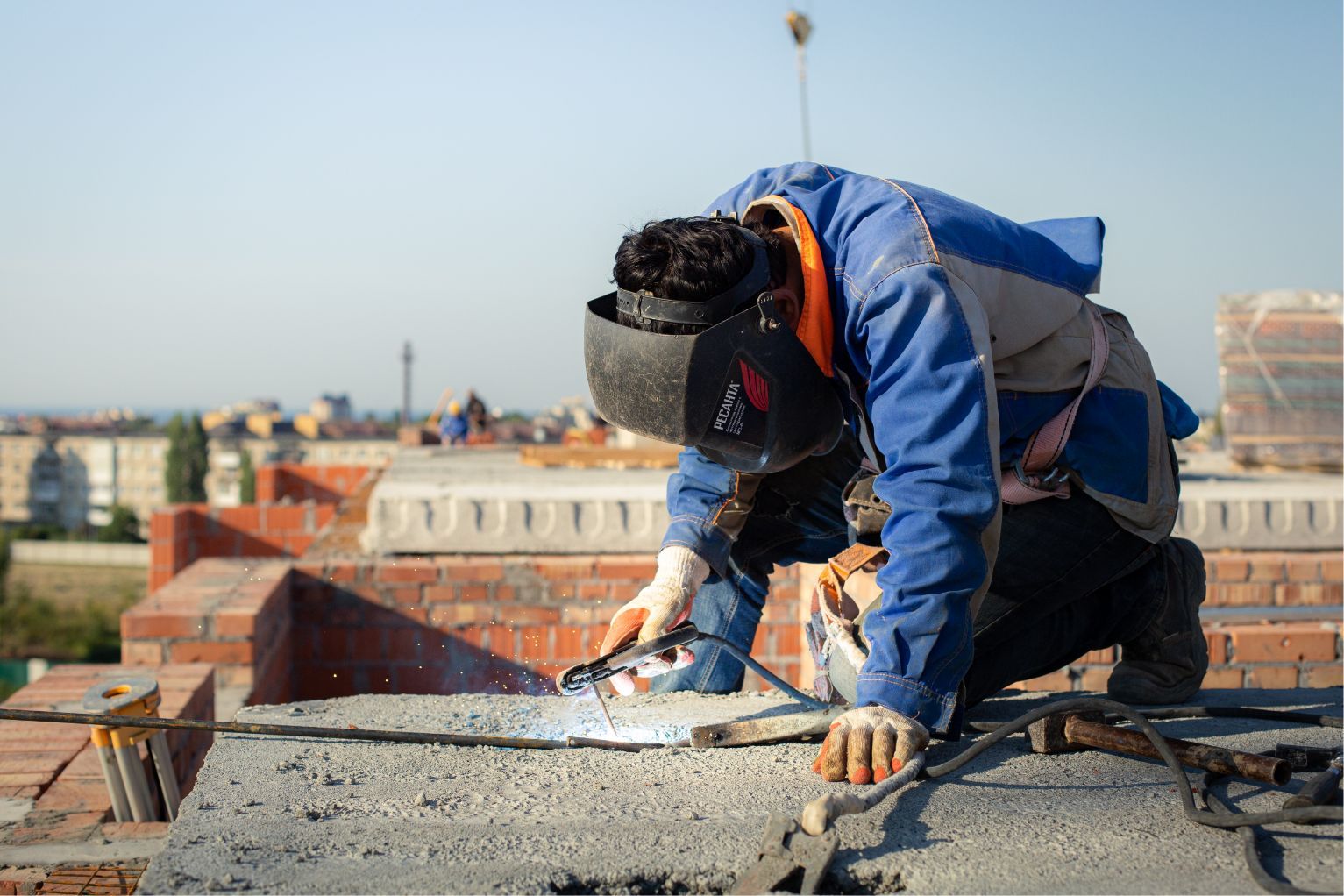 Worker welding on a rooftop, wearing safety gear amid construction materials.