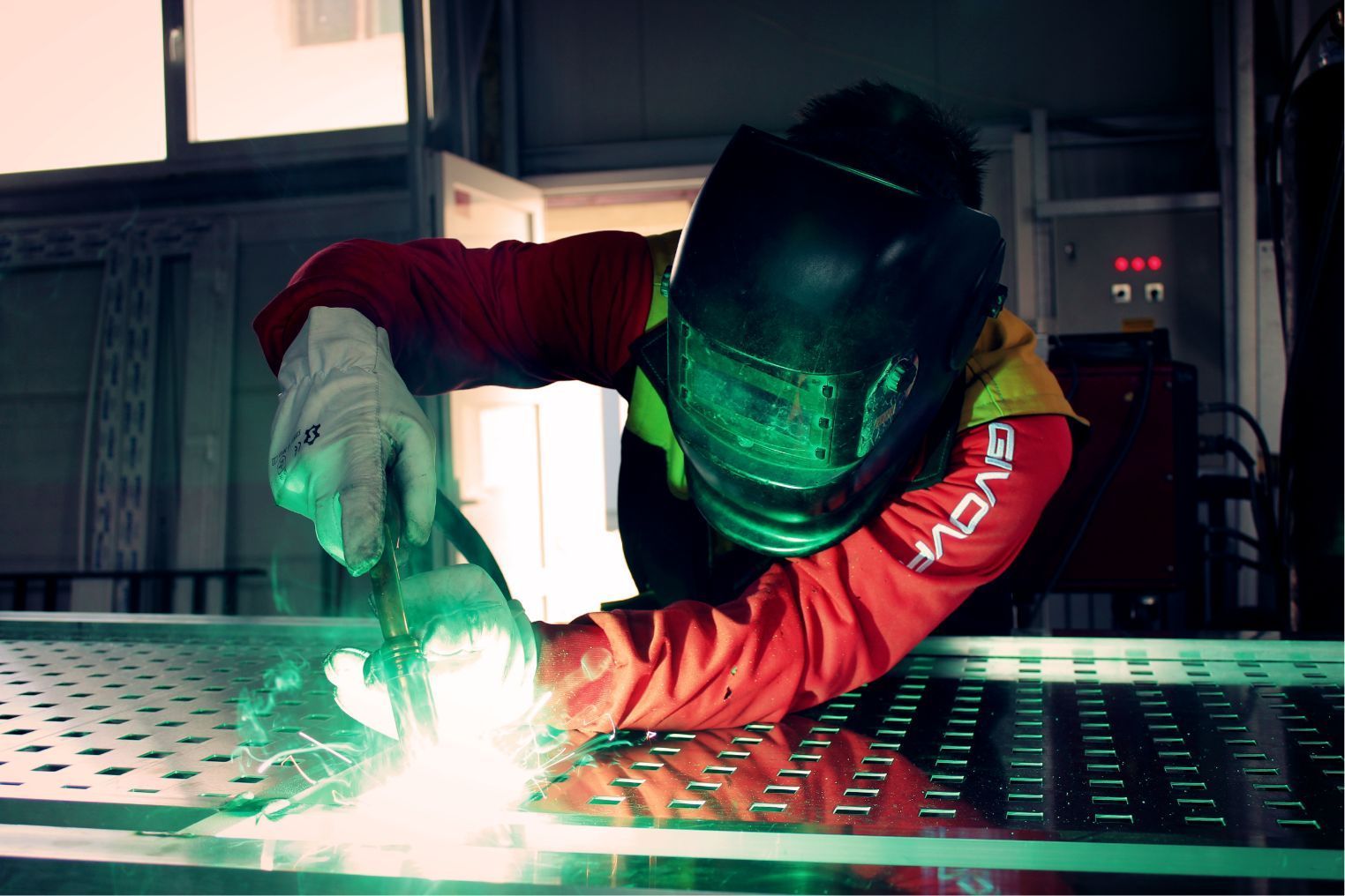 Welder in red jacket and green face shield welding at a metal workbench, sparks flying