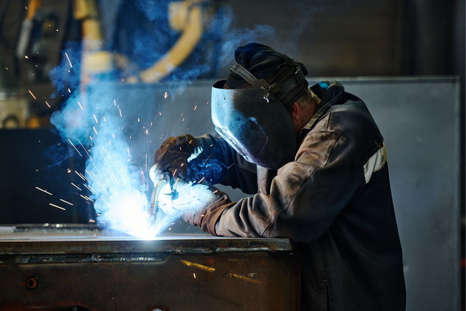 Welder in dark protective gear sparks bright blue light while working on metal in a workshop