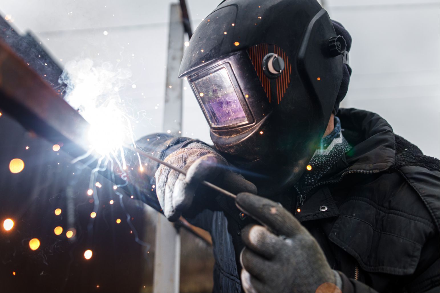 Welder in a dark helmet and gloves welding metal, bright sparks flying in an outdoor setting
