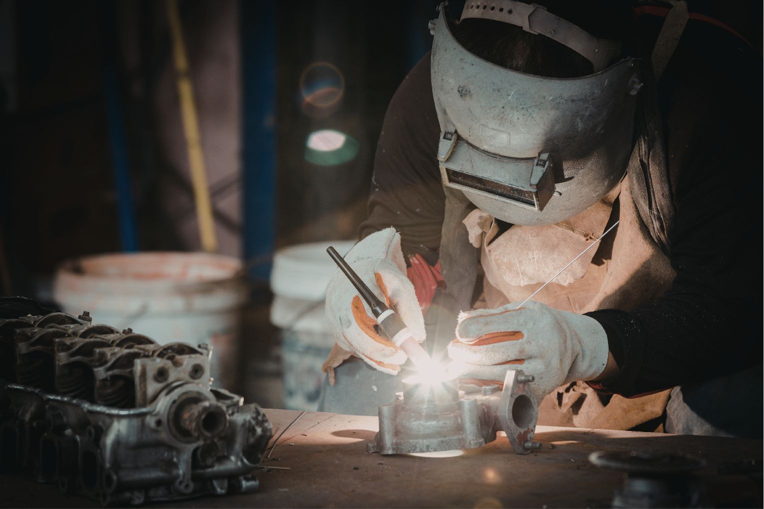 Welder in protective mask welding metal parts at a workbench in a dim workshop