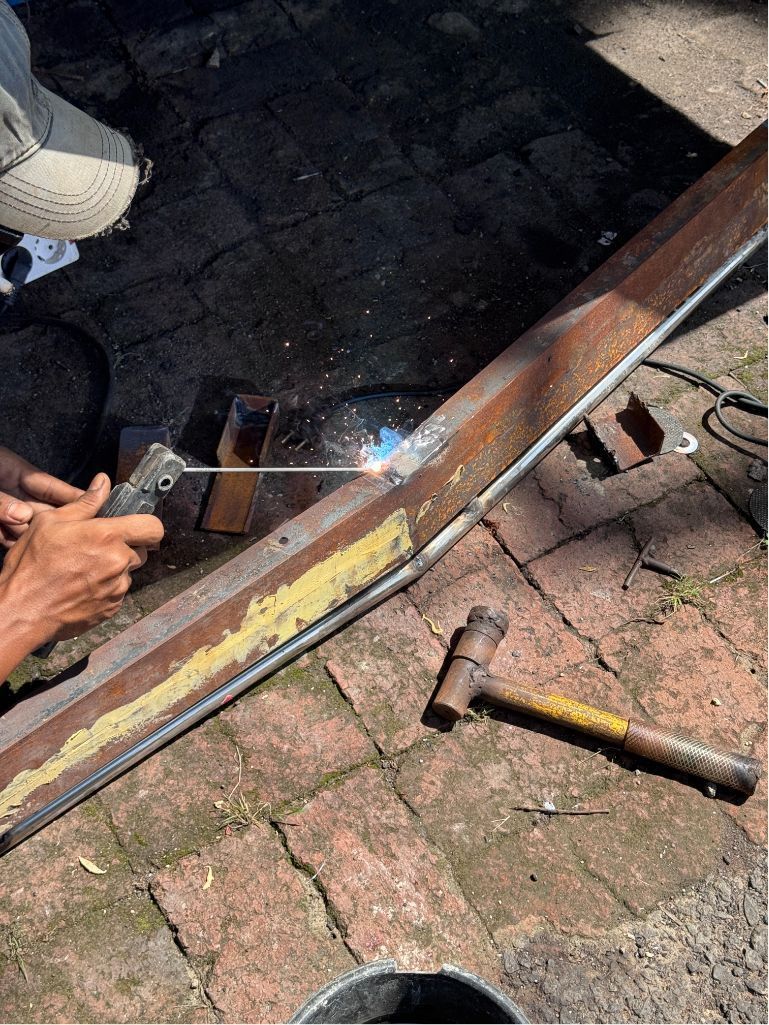 Person welding a metal frame on a brick floor, with tools nearby and bright sparks visible.