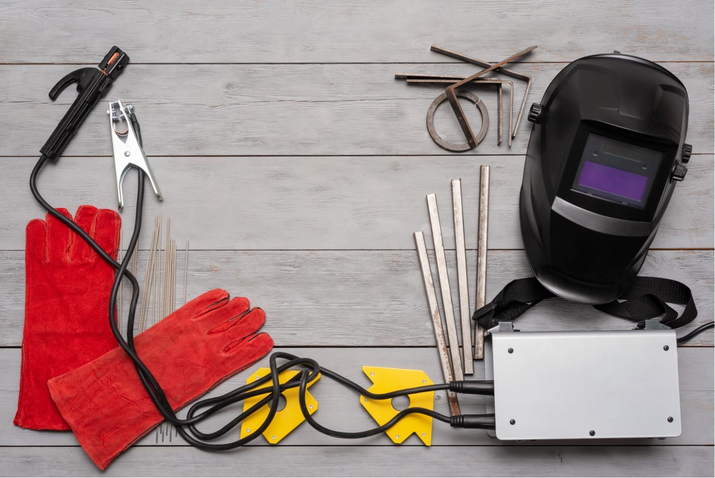 Welding tools and safety gear laid out on a gray wooden surface, including gloves, helmet, pliers, and cables