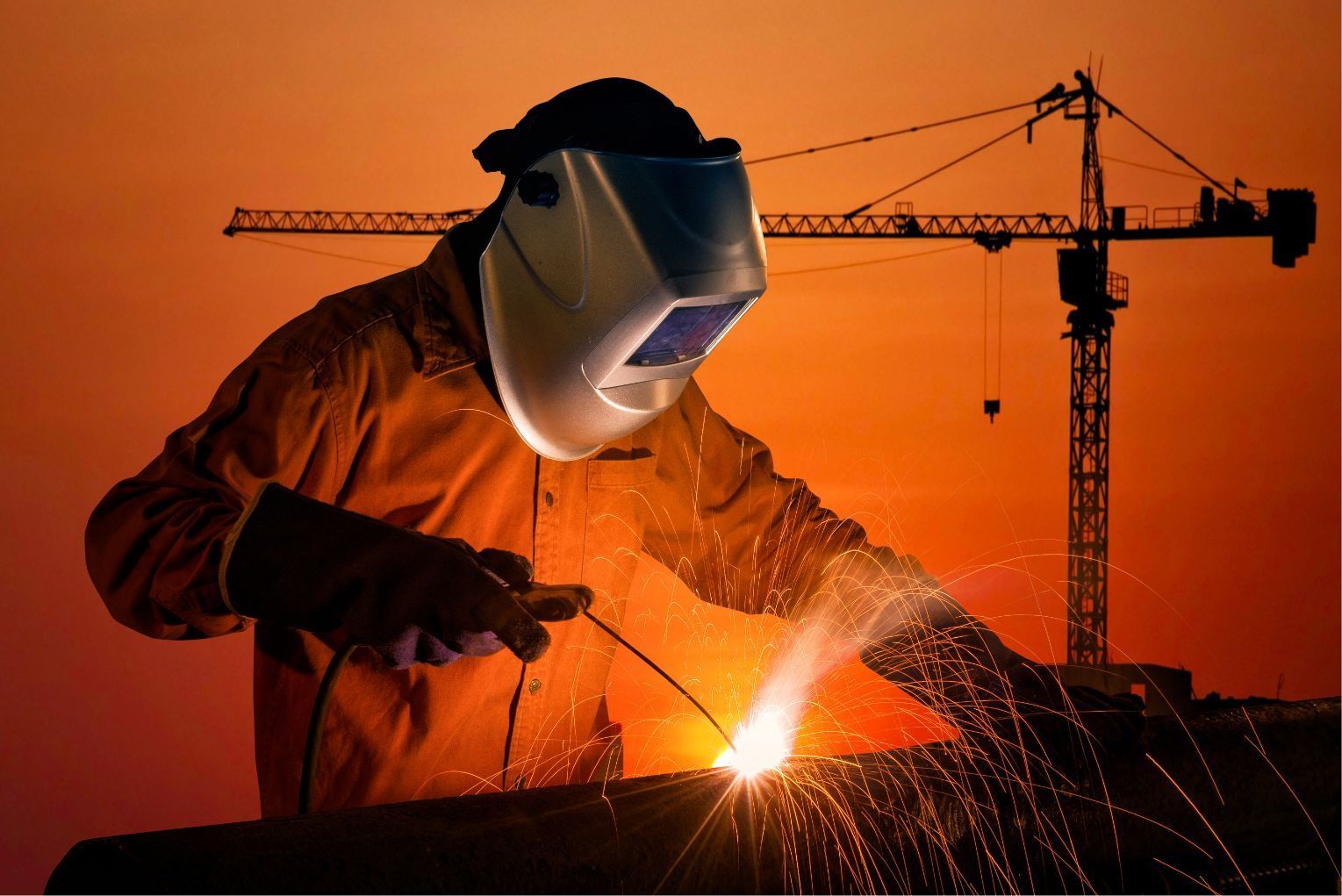Welder in protective gear sparks metal at sunset with a crane silhouetted behind him