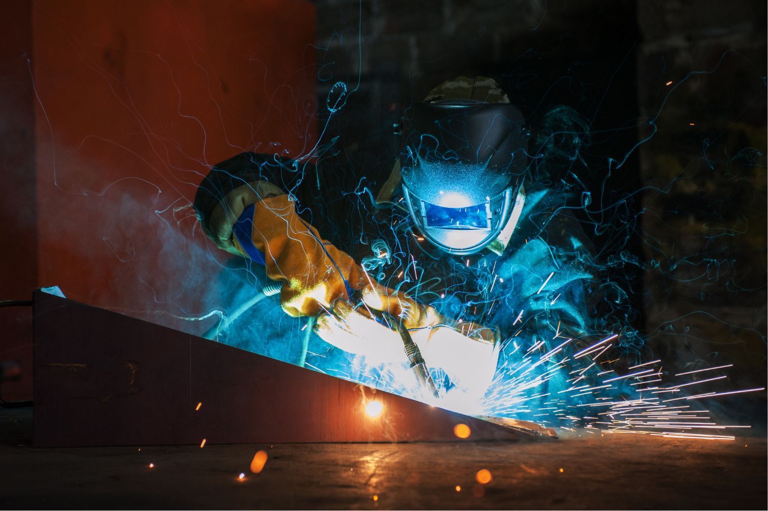Welder in blue helmet sparks metal in dark workshop with orange gloves