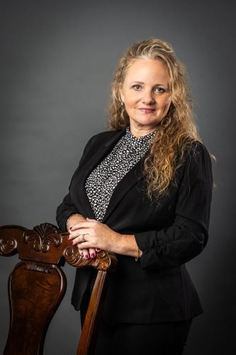 Woman in blazer leans on ornate wooden chair, smiling at the camera.