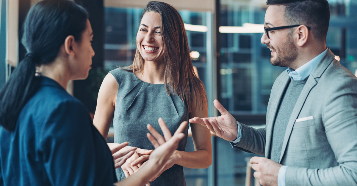 Three people in business attire have an engaging conversation, laughing and gesturing in a modern, professional office.