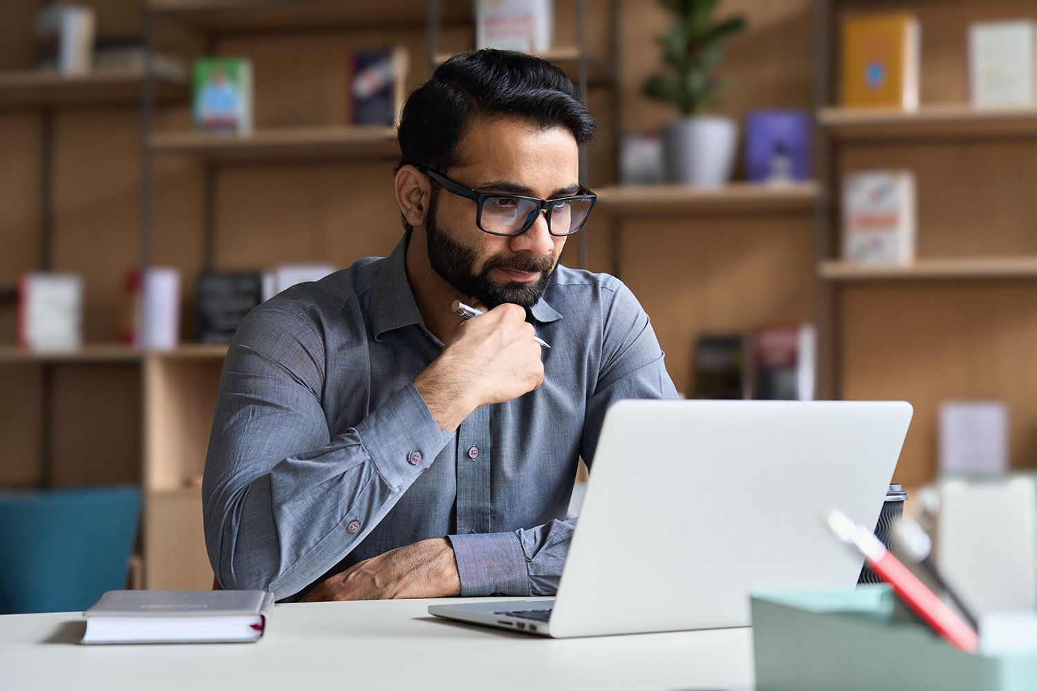 A person with a beard and glasses sits at a desk with a laptop, looking thoughtful while resting their chin on their hand.