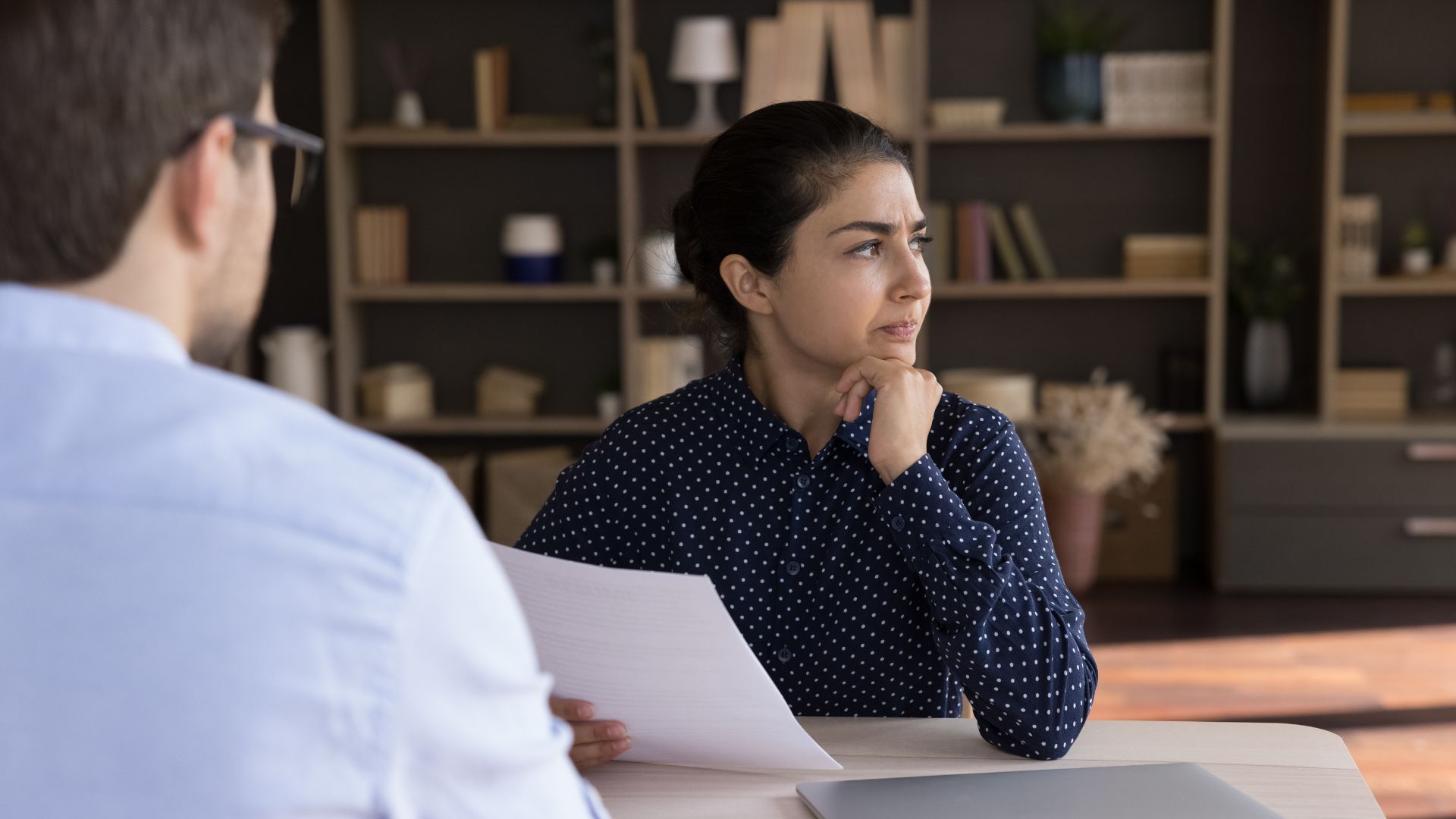A person in a dark polka-dot shirt sits at a desk holding a paper, appearing thoughtful while facing another person.