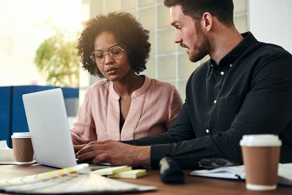 Two professionals collaborating at a desk, looking at a laptop together in a bright office.