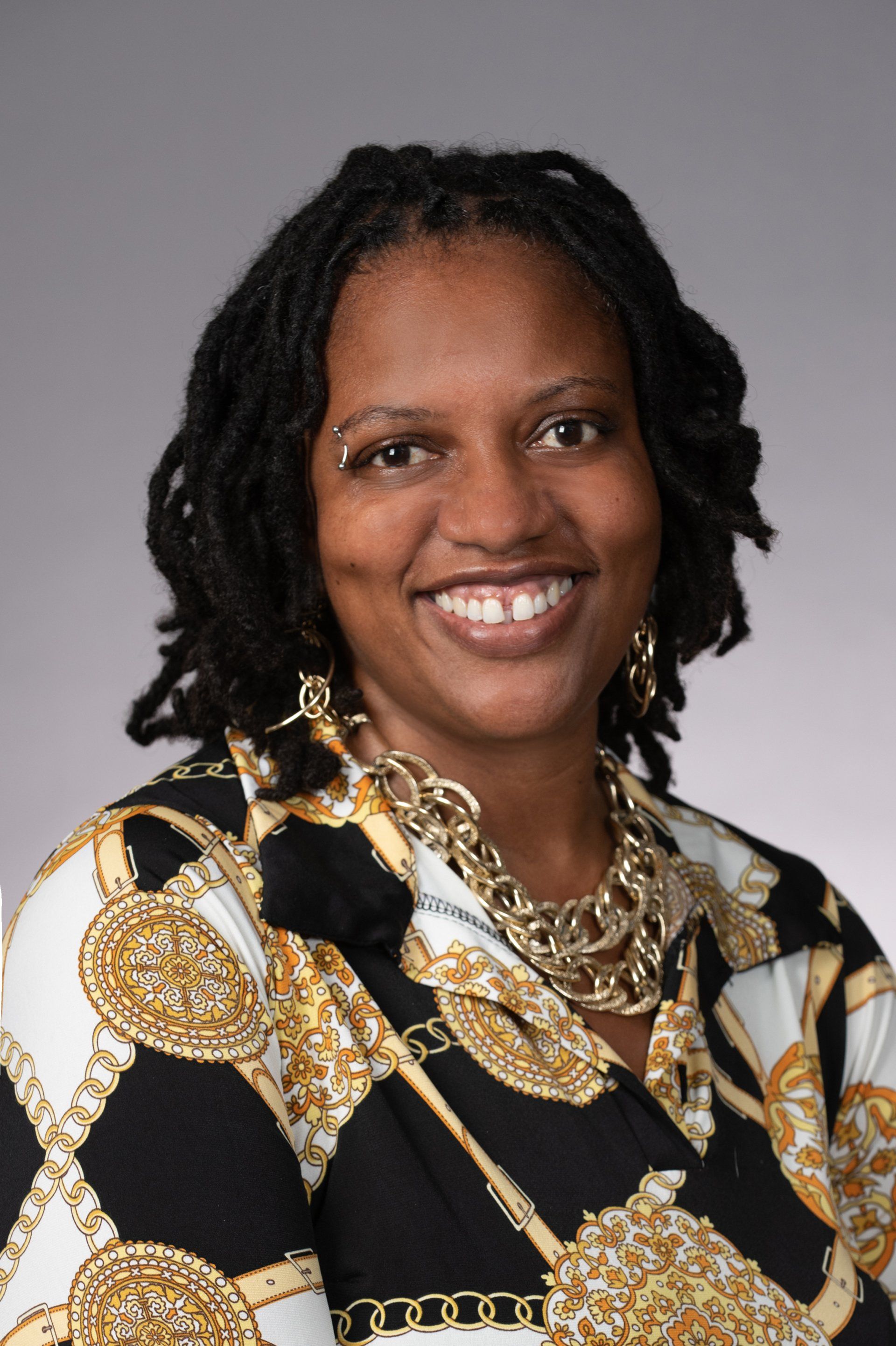 A woman with dreadlocks is wearing a black and gold shirt and a gold necklace.