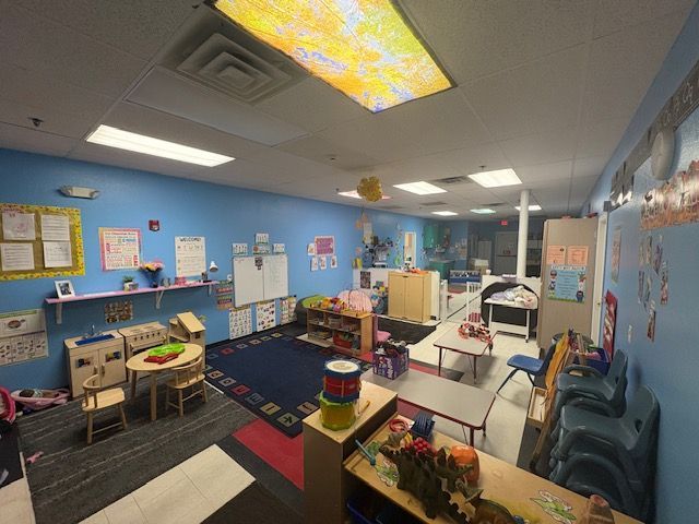 Childcare classroom: blue walls, colorful decorations, tables, chairs, and shelves filled with toys and supplies.