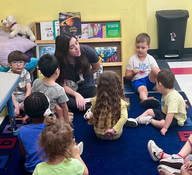 Teacher reading to a group of children seated on a blue rug in a classroom, with books on a shelf in the background.