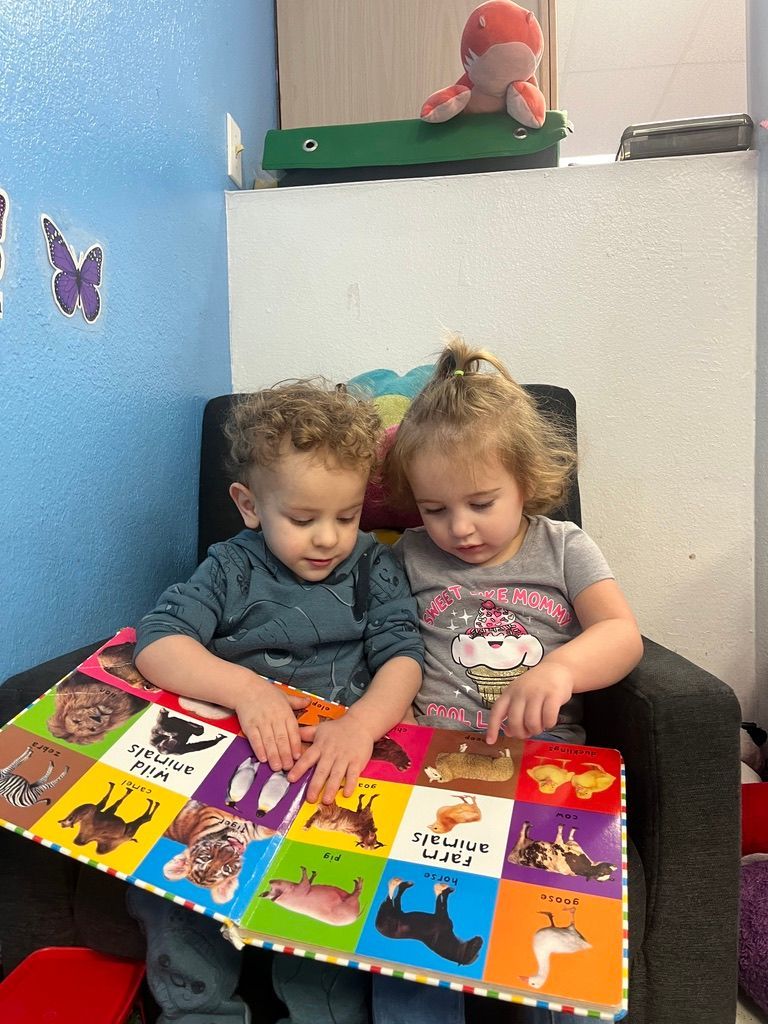 Two toddlers looking at a colorful book in a room with blue walls, a stuffed octopus, and a butterfly sticker.