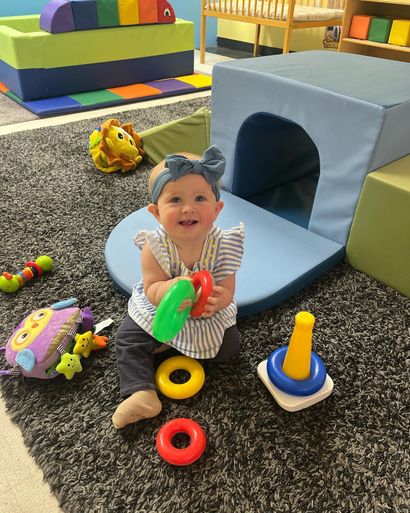 Smiling baby plays with colorful toys in a playroom; blue blocks and gray carpet surround her.