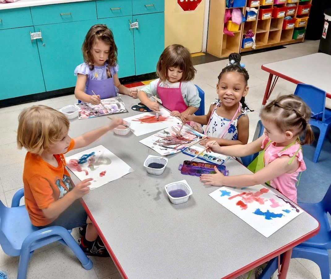 Children painting at a table in a classroom. They are using colorful paint and smiling.