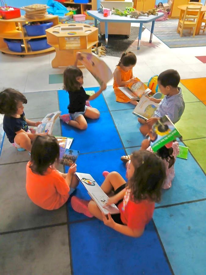Children seated in a circle, reading books in a classroom with colorful mats and tables.