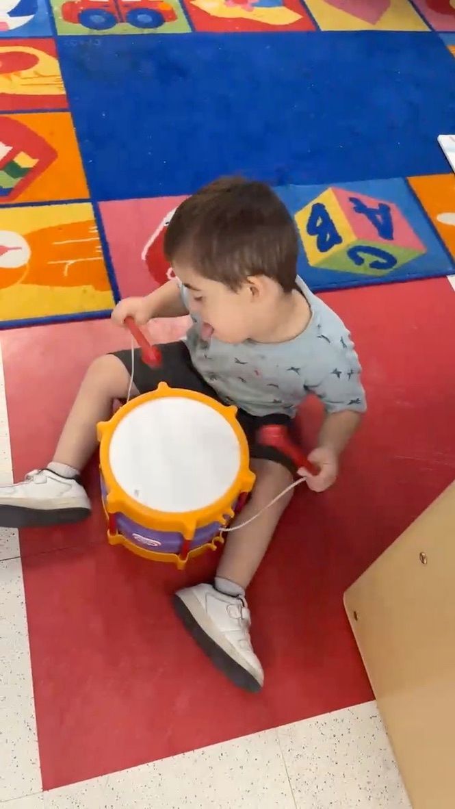 Child sitting on red mat, playing a drum with drumsticks on colorful floor.