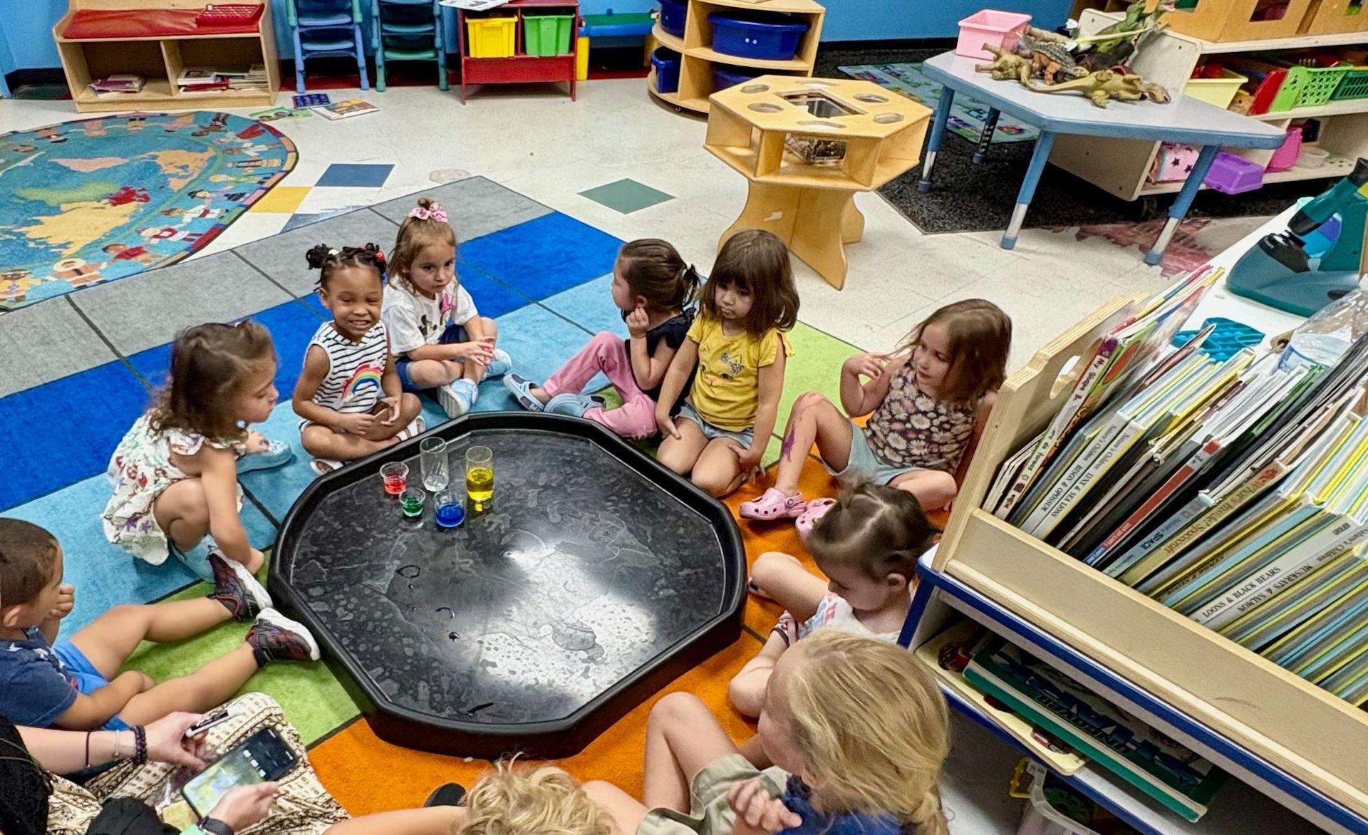 Children sit around a black tray with toys, inside a colorful classroom.