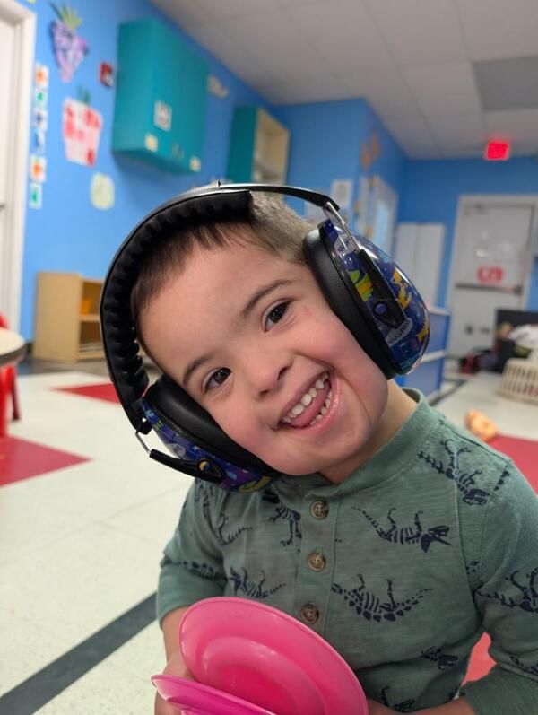 Child with Down syndrome wearing noise-canceling headphones, smiling in a room with colorful decor, holding a pink toy.