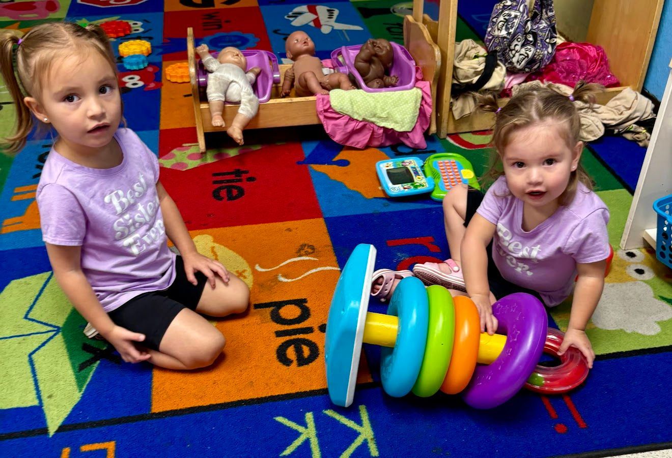 Two young children playing with a ring stacker toy on a colorful rug. Dolls and a toy crib are in the background.