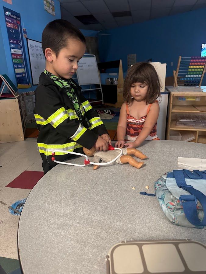 Child in firefighter jacket uses a stethoscope on doll. Another child watches at a table. Classroom setting.