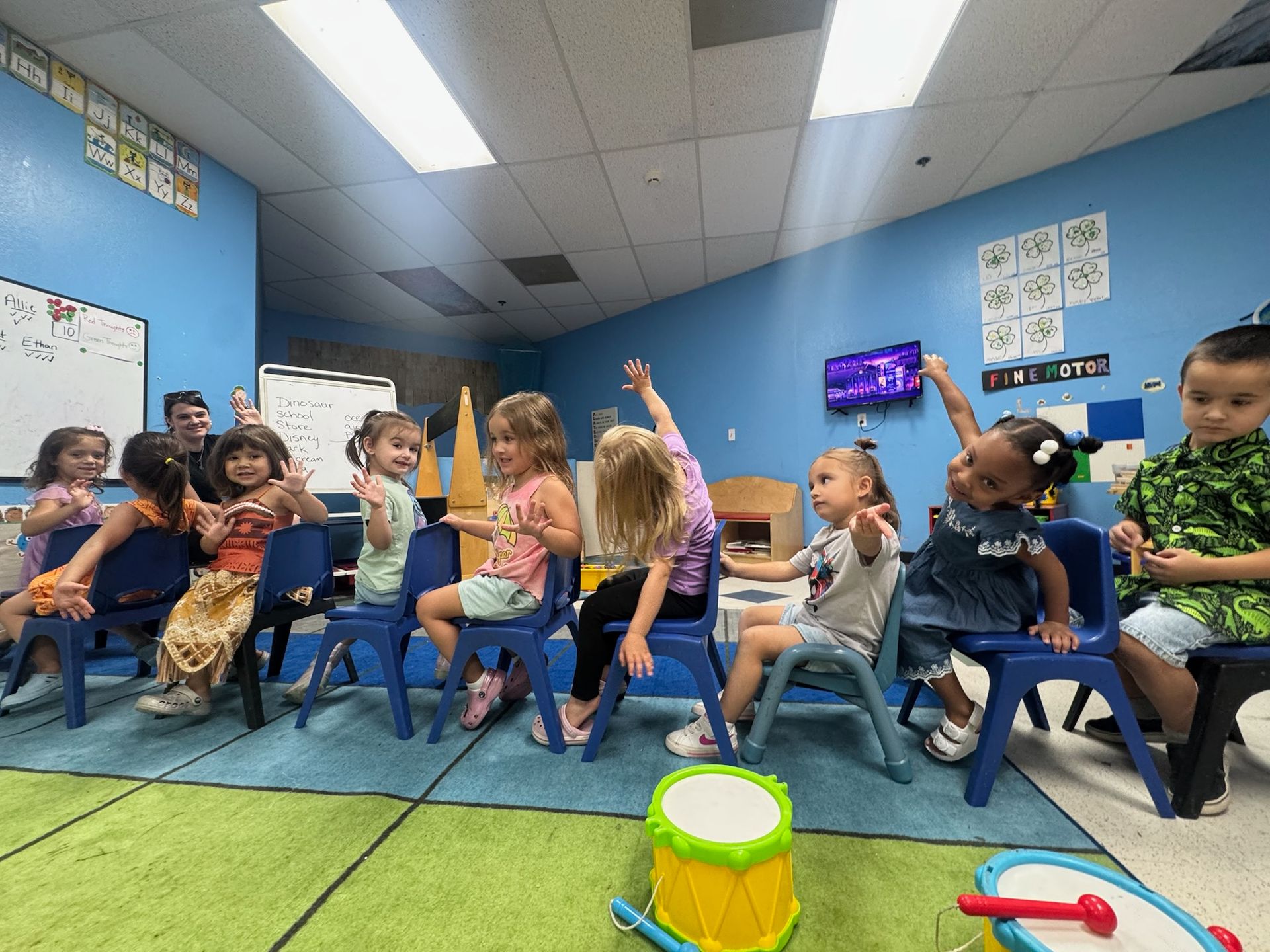 Children in a classroom seated in chairs, raising their hands with a teacher.